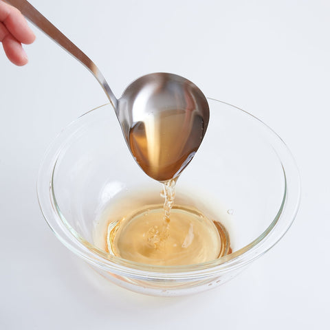 a hand pours soup broth into a clear glass bowl using a left-handed soup ladle