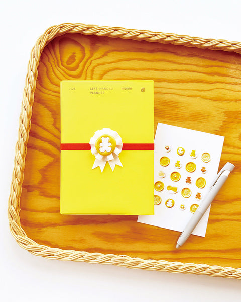 2025 Hidari Left-Handed Planner in yellow with decorative ribbon and stickers on a wooden tray, featuring a pen nearby.