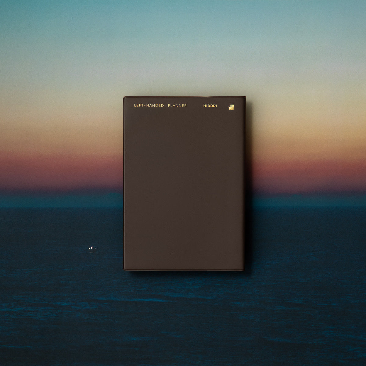 Hidari Left-Handed Planner with dark cover against a serene sunset background, showcasing the elegant design.